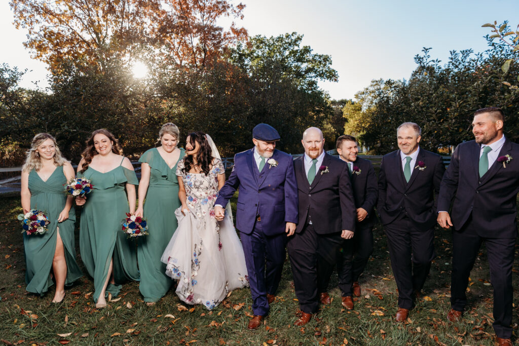 Outdoor autumn wedding party walking together: bridesmaids in green dresses, a bride in a white gown, and groomsmen in suits.