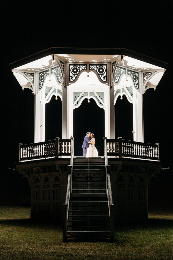 Bride and groom standing on a lit, ornate white gazebo at night.