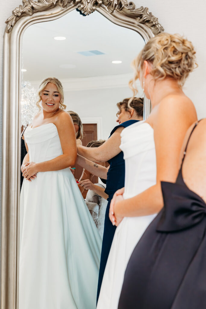 A smiling woman in a pale blue formal gown stands before an ornate mirror as friends help her adjust dresses.