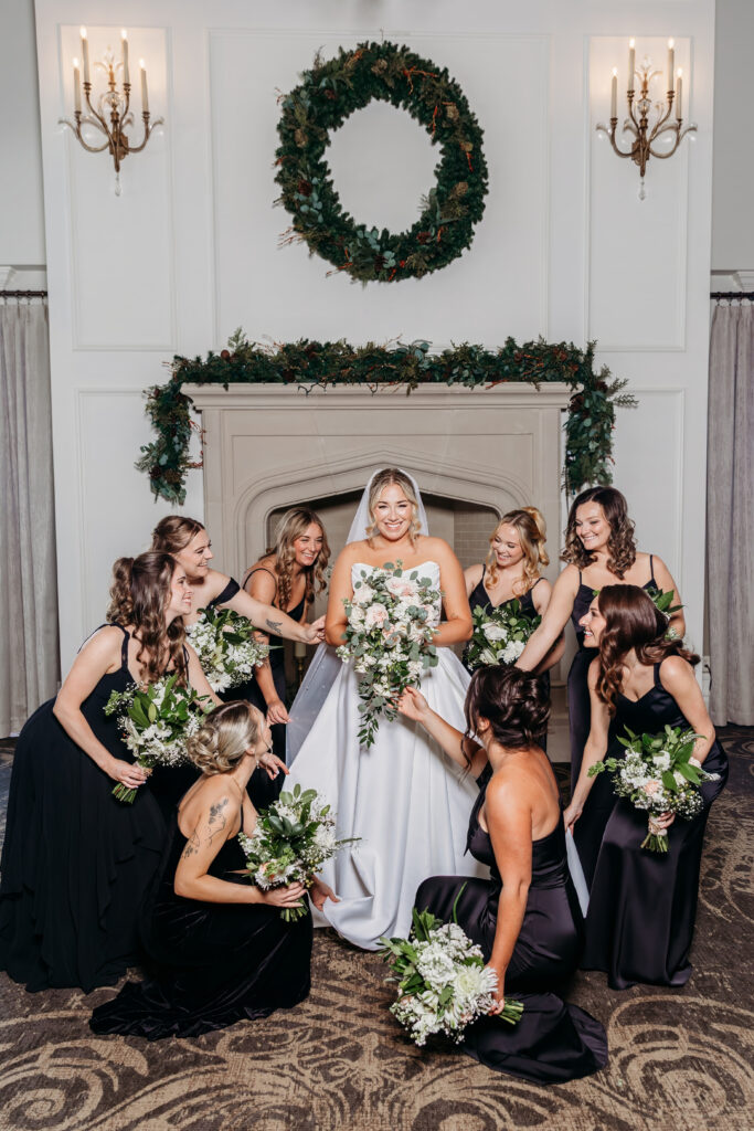Bride in a white wedding gown stands center with bridesmaids in black dresses, all holding bouquets, in a room decorated with a fireplace, greenery, and a large wreath.