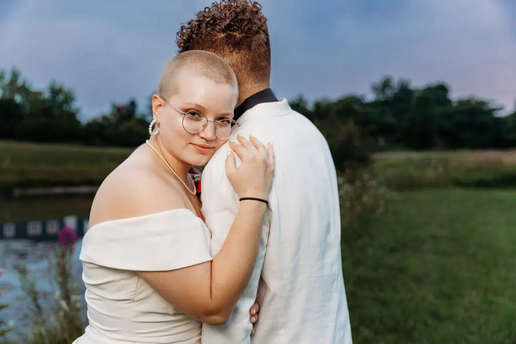 Two people embrace near a pond. One person with glasses looks at the camera while the other faces away. Trees and grassy landscape in the background.