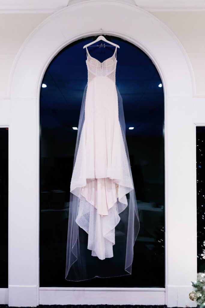 A white wedding dress hangs elegantly on a hanger, displayed in front of an arched window, with a dark background.