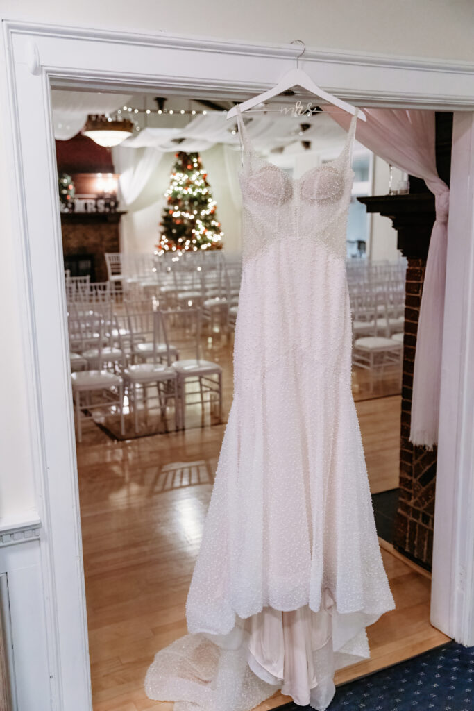A wedding dress hangs in a decorated venue with chairs and a lit Christmas tree in the background.