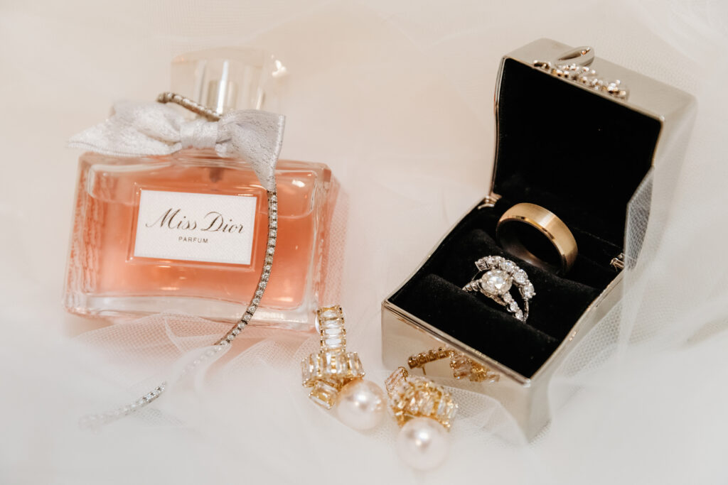 Perfume bottle, jewelry, and rings displayed elegantly on soft fabric. Includes a Miss Dior bottle with pearl earrings and ornate rings.