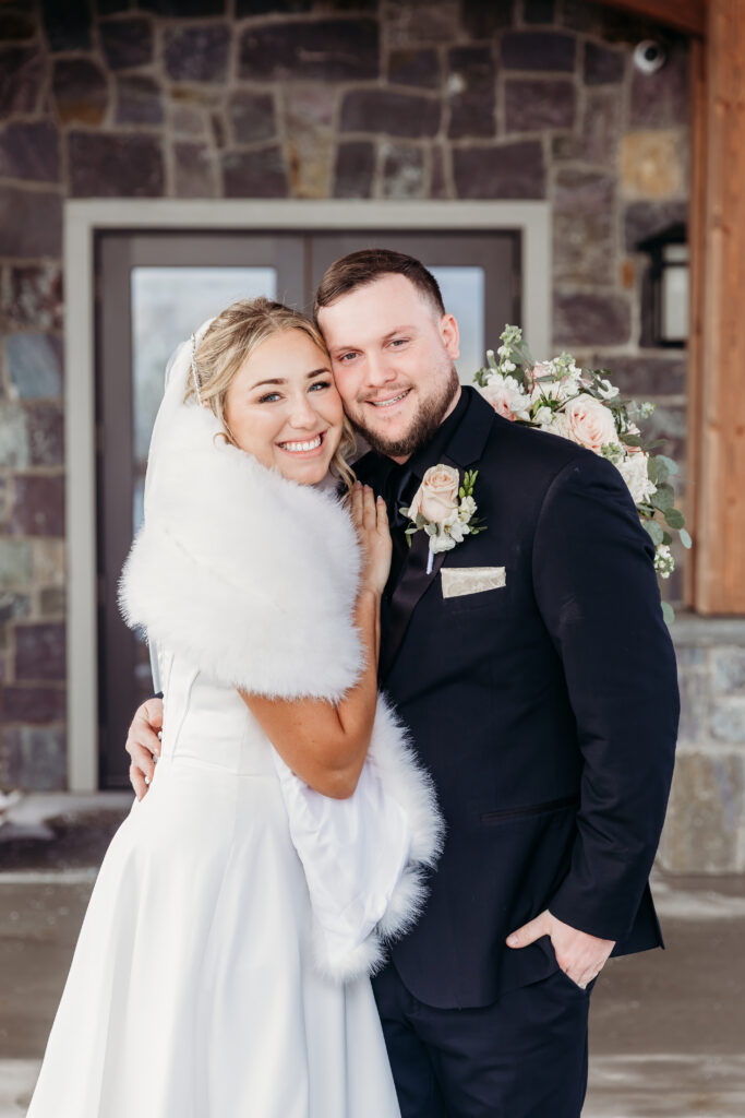 Bride in a white wedding dress with a fur wrap stands next to a groom in a dark suit outside a stone building, both smiling at the camera.