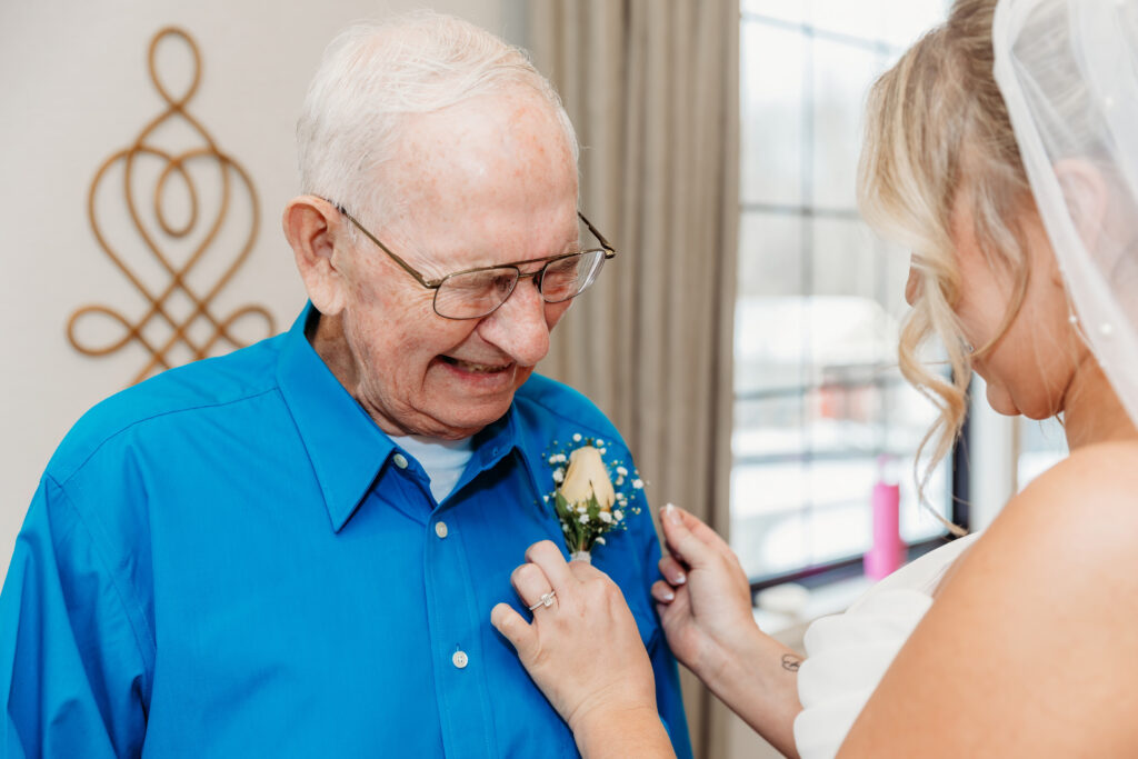An elderly man in a blue shirt smiles as a bride pins a boutonniere to his lapel.