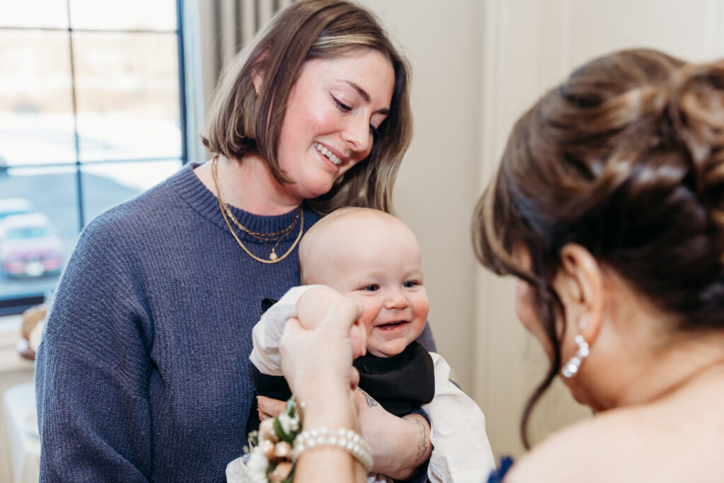 A smiling baby being held by a woman in a blue sweater, with another woman wearing a bun and pearl earrings looking on.