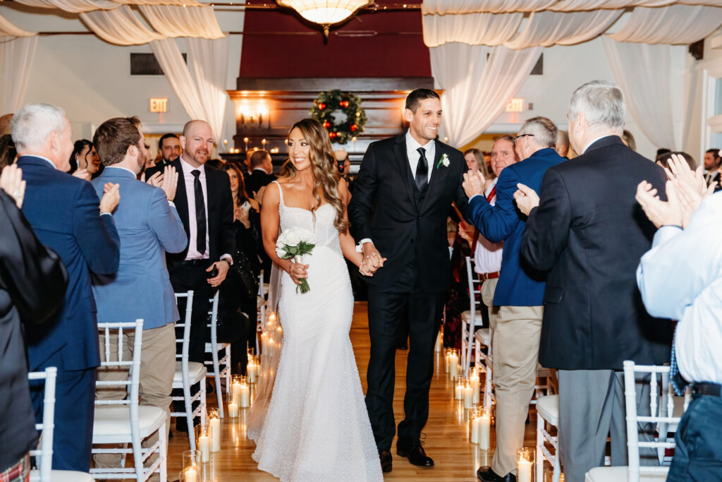 A newlywed couple walks down a candlelit aisle, holding hands, surrounded by applauding guests in a decorated indoor venue.