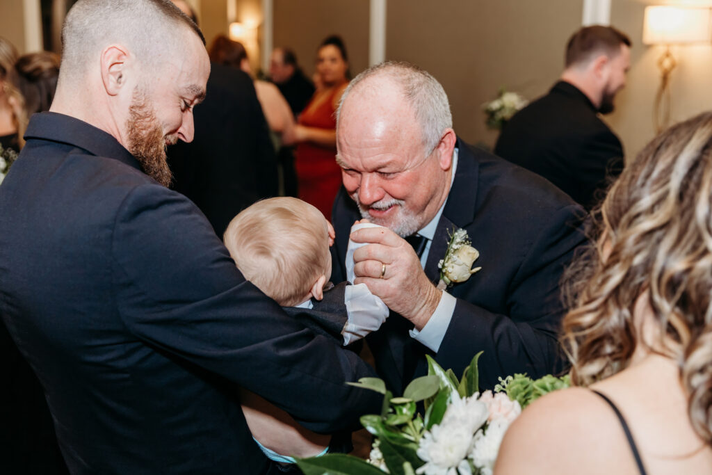 An elderly man with a boutonniere smiles as he shakes hands with a baby held by a man at a wedding reception.