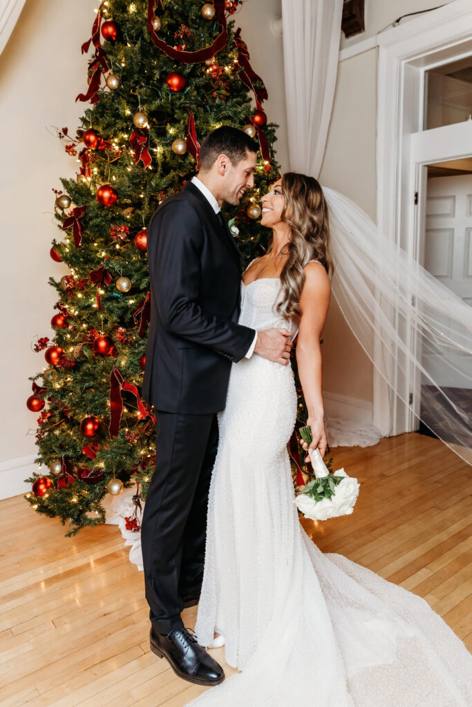 Two people dressed formally, standing by a decorated Christmas tree. They are indoors, smiling, and holding a bouquet of white flowers.