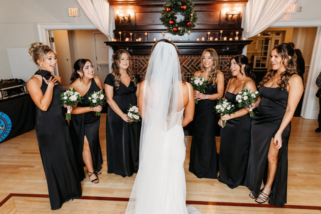 A person in a wedding dress faces six people in black dresses holding bouquets, standing in front of a festive fireplace indoors.