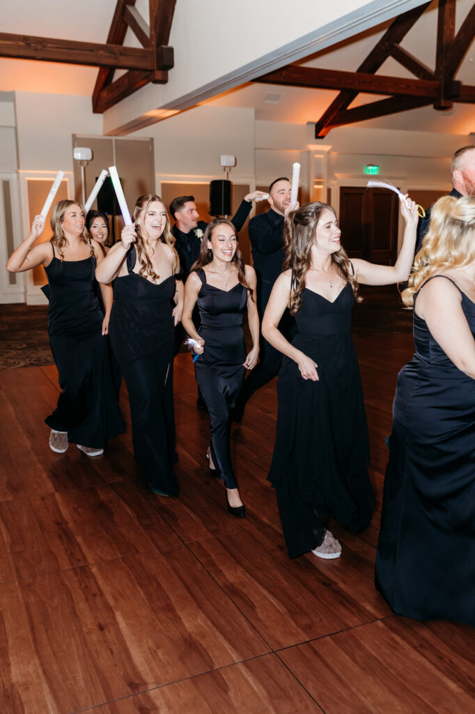 Group of women in black dresses walking in a line on a wooden floor in an indoor event space.