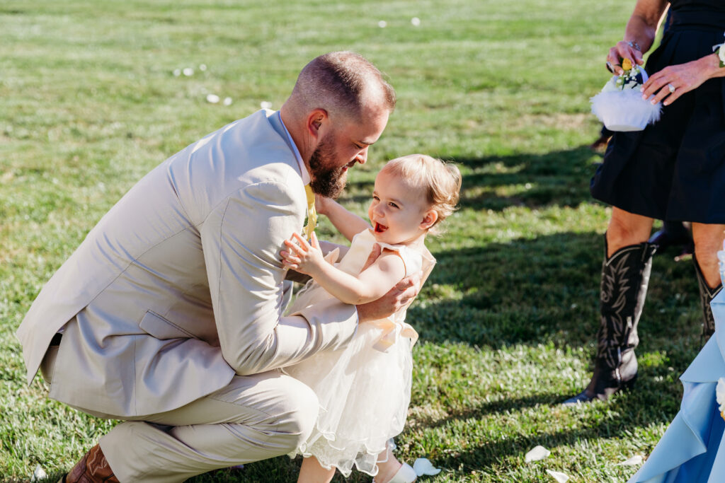 A man in a light suit kneels on the grass and interacts with a young girl in a white dress outdoors.