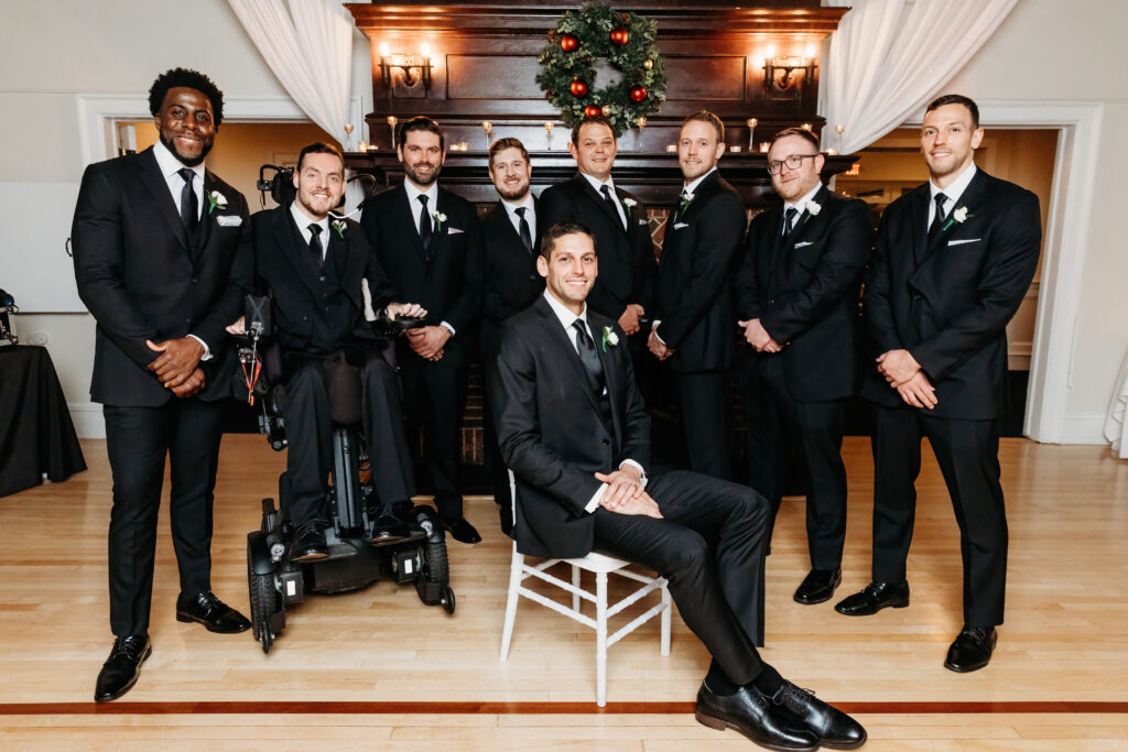 Nine people in formal black suits pose indoors by a decorated fireplace, one person seated in a wheelchair, another on a chair.