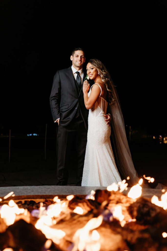 Two people dressed formally, standing by a fire at night, with a dark background and subtle lights in the distance.