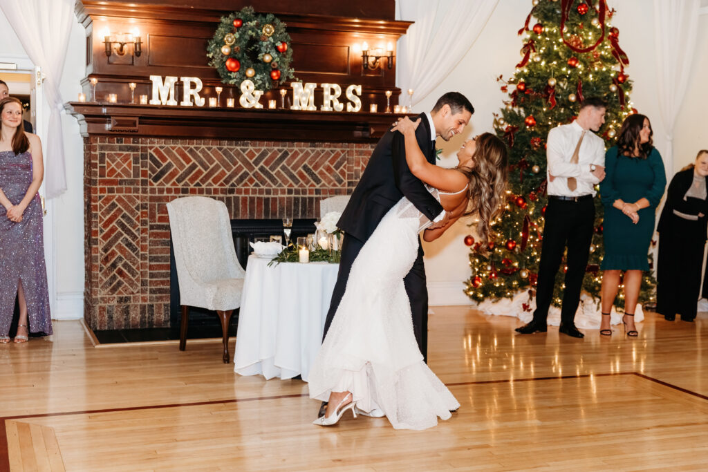 A person in a wedding dress and another in formal wear dance near a decorated fireplace and Christmas tree, observed by others.