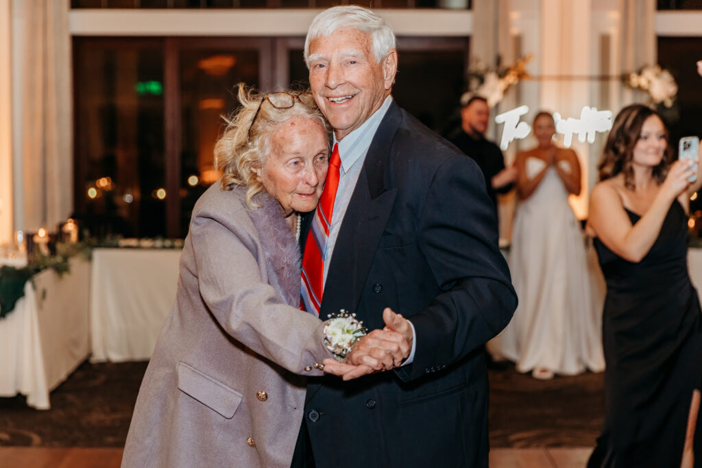 An elderly couple in formal attire embraces and smiles at a reception, with the woman wearing a lavender coat and the man in a dark suit with a red tie.