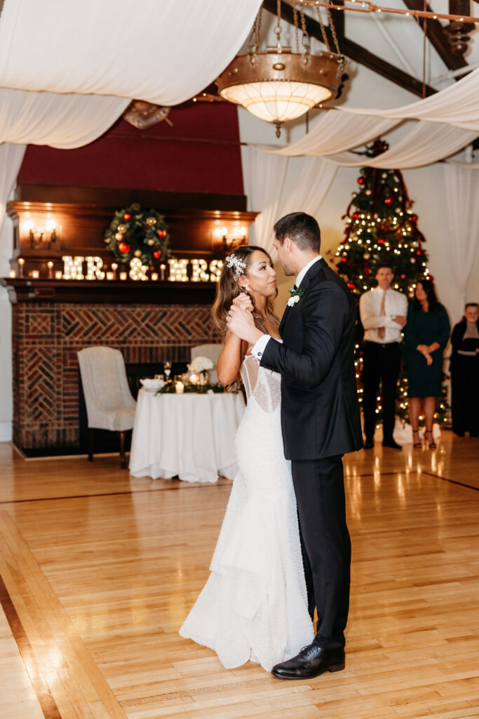 A person in formal attire dances with another in a decorated room, featuring a fireplace and festive Christmas tree, under elegant drapery.