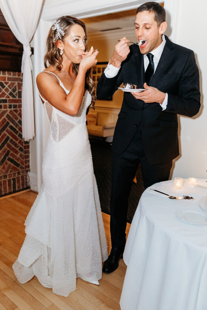 Two people in formal attire enjoy dessert at a reception. They stand near a table with candles in a warmly decorated room.