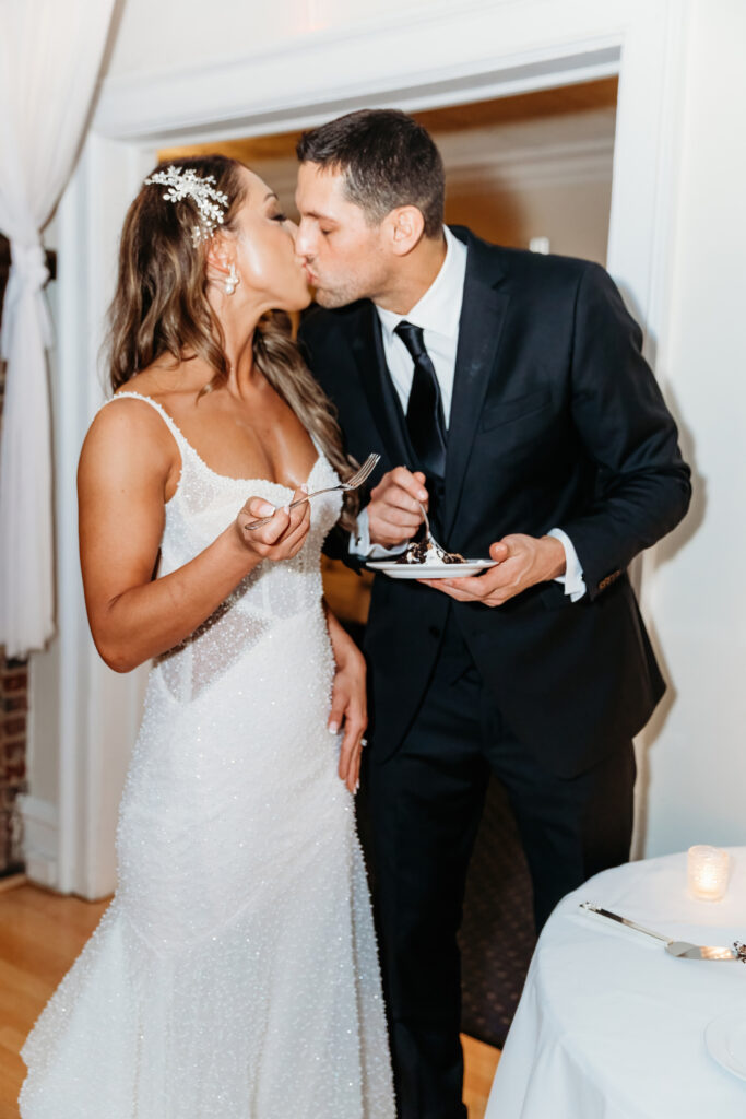 Two people in formal attire share a kiss while holding forks and cake plates, captured in a warmly lit room.