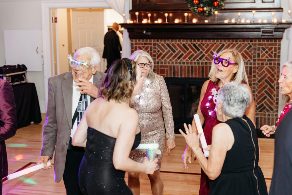 People celebrating indoors, wearing colorful glasses and party attire near a brick fireplace decorated with candles and a wreath.