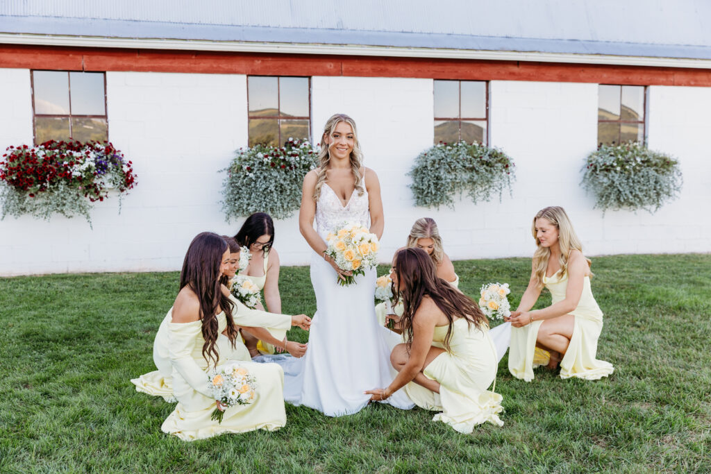 Bride in a white wedding gown stands on a grassy lawn as bridesmaids in pale yellow dresses kneel around her, holding bouquets, outside a white building with window boxes.
