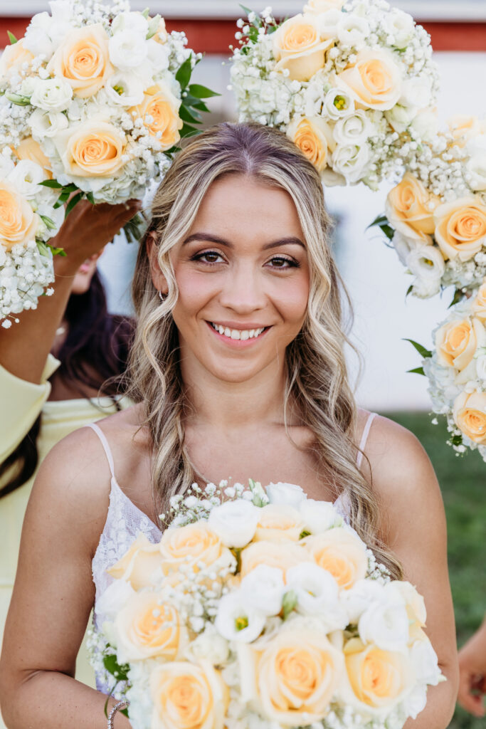 Smiling bride with braided hair holding a bouquet of white and peach roses, standing beneath floral arches.