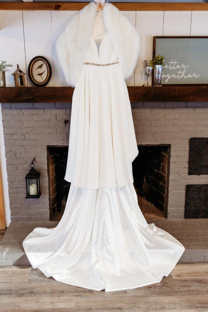 A white wedding dress with a fur shawl on a hanger, displayed in front of a rustic brick fireplace with decor.