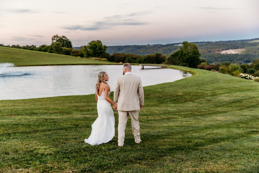 A bride in a white wedding dress and a groom in a light suit stand on a grassy hill overlooking a pond and rolling countryside.