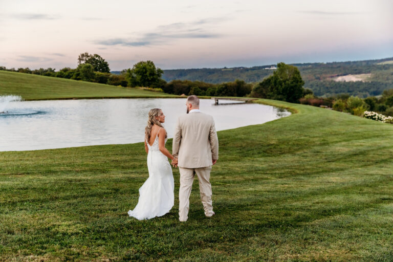 A bride in a white wedding dress and a groom in a light suit stand on a grassy hill overlooking a pond and rolling countryside.