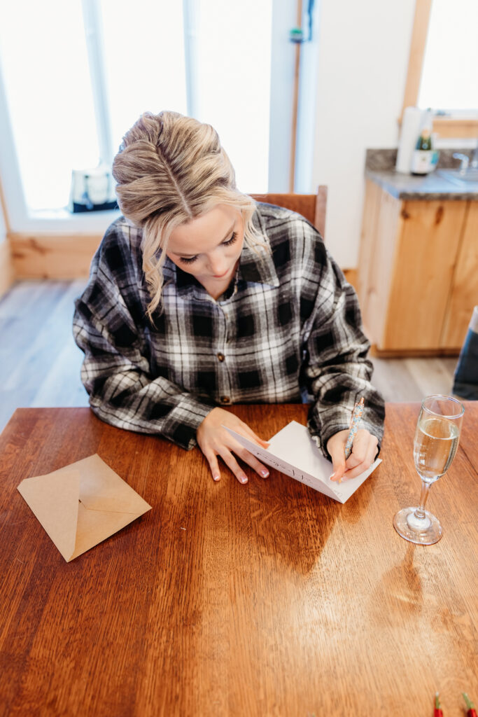 A person with blonde hair writes a note at a wooden table, next to a brown envelope and a glass of champagne.