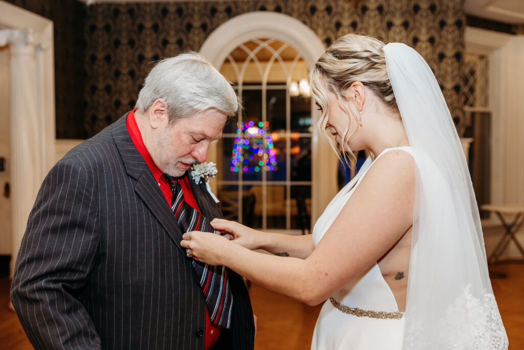 A person in a wedding dress adjusts another person's tie in an elegant room with an arched window and festive lights.
