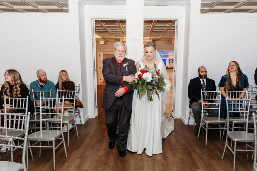 A person escorts another holding a bouquet down an aisle, while seated guests look on in a modern indoor setting.