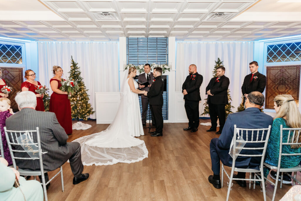 A wedding ceremony with people in formal attire, standing on a decorated indoor venue with white drapes, Christmas trees, and guests seated on chairs.