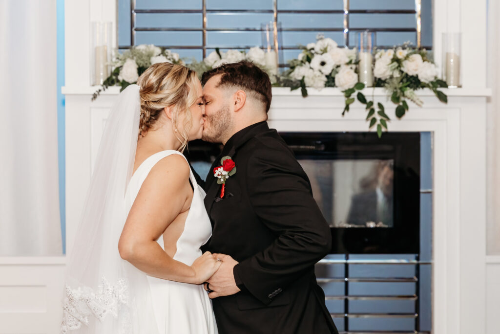 Two people kiss in front of a decorative mirror, adorned with flowers, during a special occasion or ceremony.