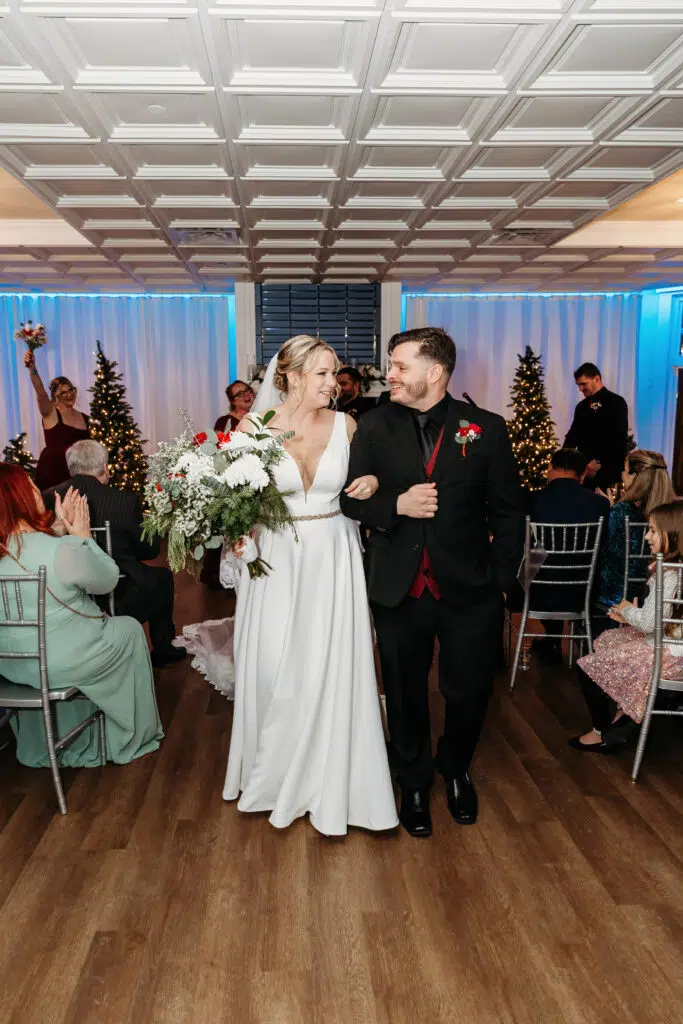 A person in a white dress and another in a dark suit walk arm-in-arm indoors, surrounded by guests and festive decorations.