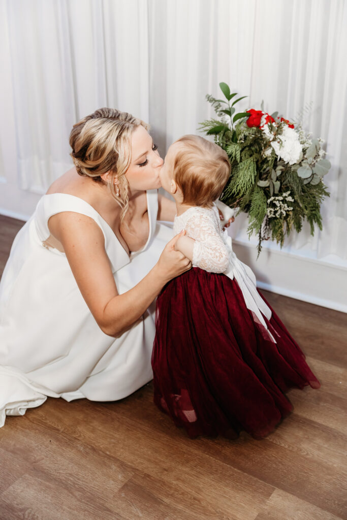 A person in a white dress kisses a young child in a red dress, with a bouquet featuring red and white flowers nearby.
