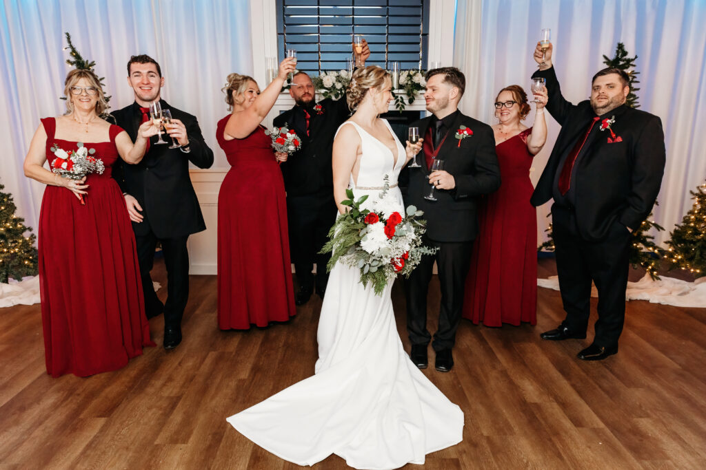A wedding party celebrates with champagne toasts. The couple stands at the center, surrounded by six people in formal attire, holding bouquets.