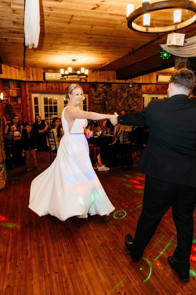 A person in a white dress dances with another in a suit in a warmly lit wooden venue with guests observing.