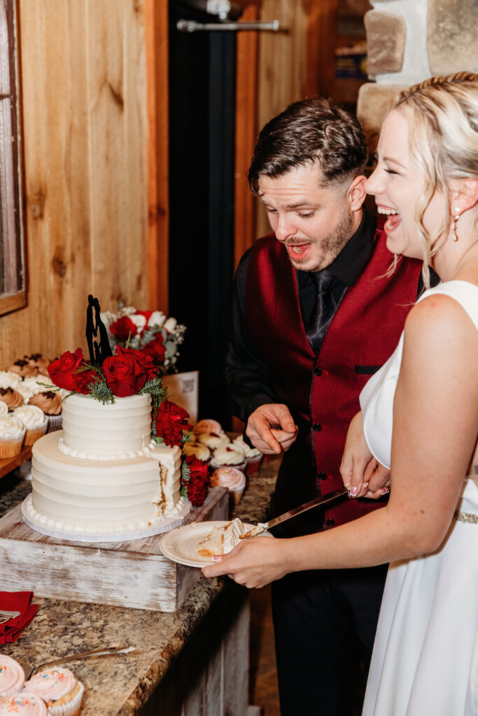 Two people smiling while cutting a white cake with red roses in a cozy, rustic setting. Several cupcakes are displayed nearby.