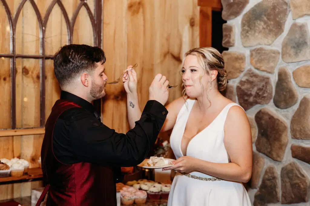 Two people in formal attire share cake in a cozy setting with rustic wooden and stone decor, surrounded by cupcakes and soft lighting.