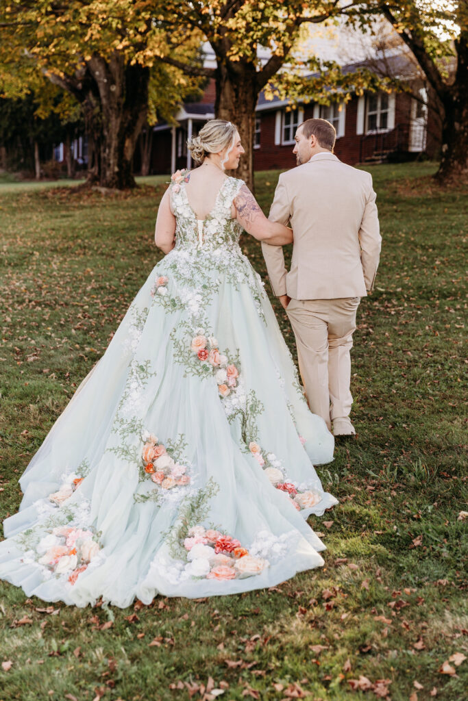 Back view of a bride in a pale blue floral wedding gown with a long train walking beside a groom in a light tan suit on a grassy lawn.