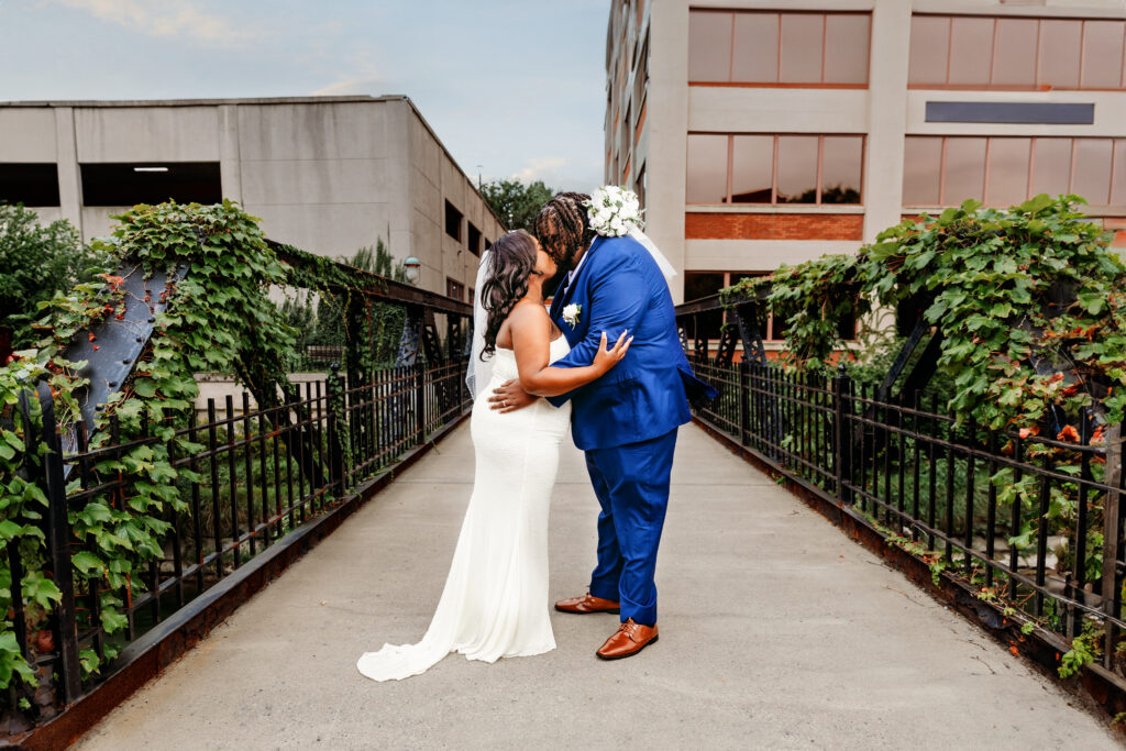 Two people in wedding attire embrace on a vine-covered bridge with modern office buildings in the background, during daytime.