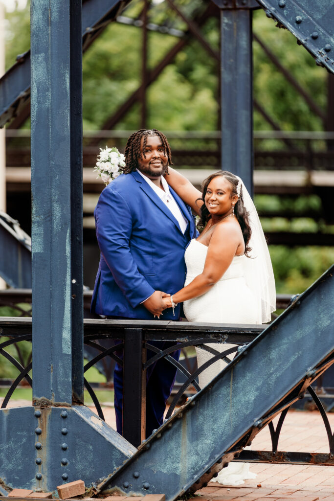 A couple in wedding attire poses for a photo on an iron bridge, surrounded by lush greenery.