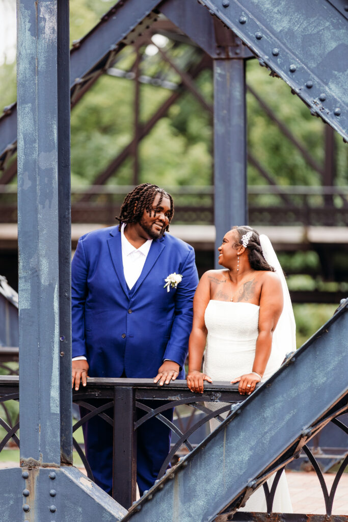 A newlywed couple stands on an old metal bridge, smiling at each other with lush greenery in the background. The person wears a blue suit.