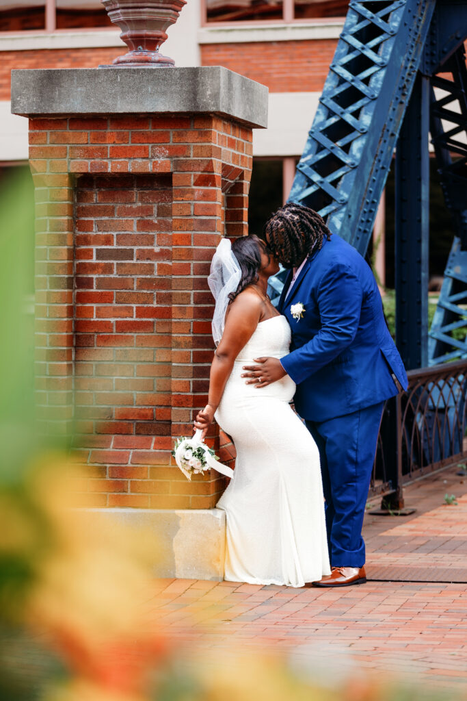 A person in a white dress and veil embraces another in a blue suit, standing near a brick pillar and metal structure.