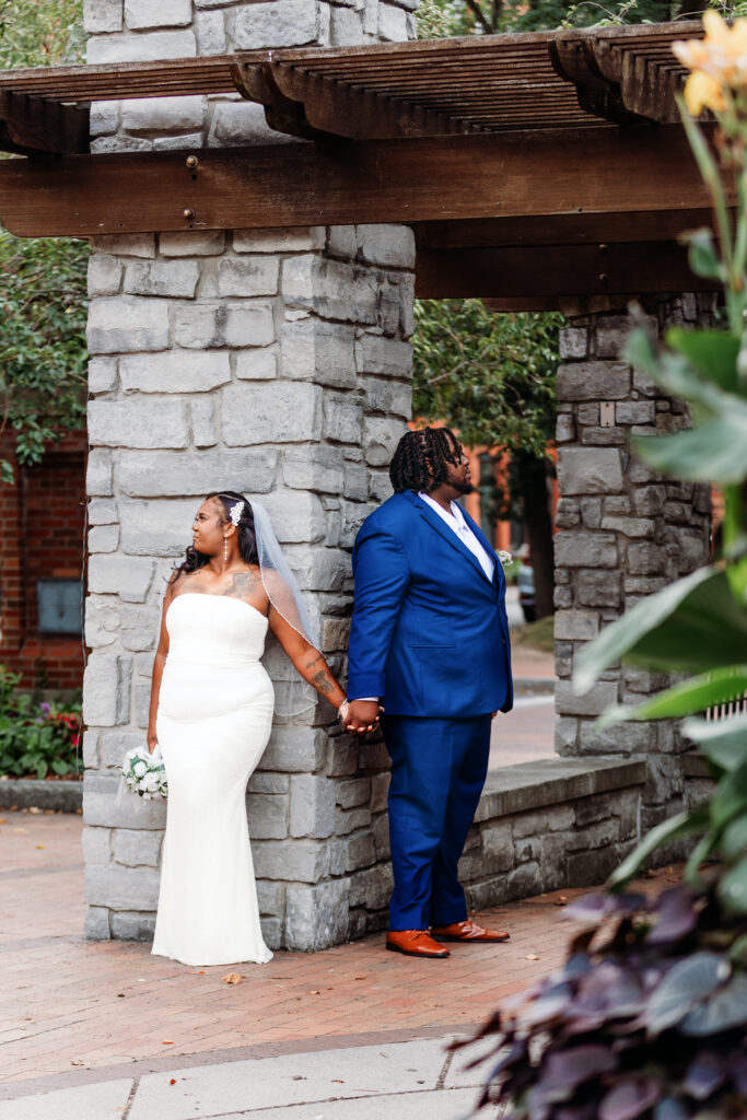 Two people in wedding attire stand back-to-back, holding hands near a stone structure with wooden beams, surrounded by greenery.