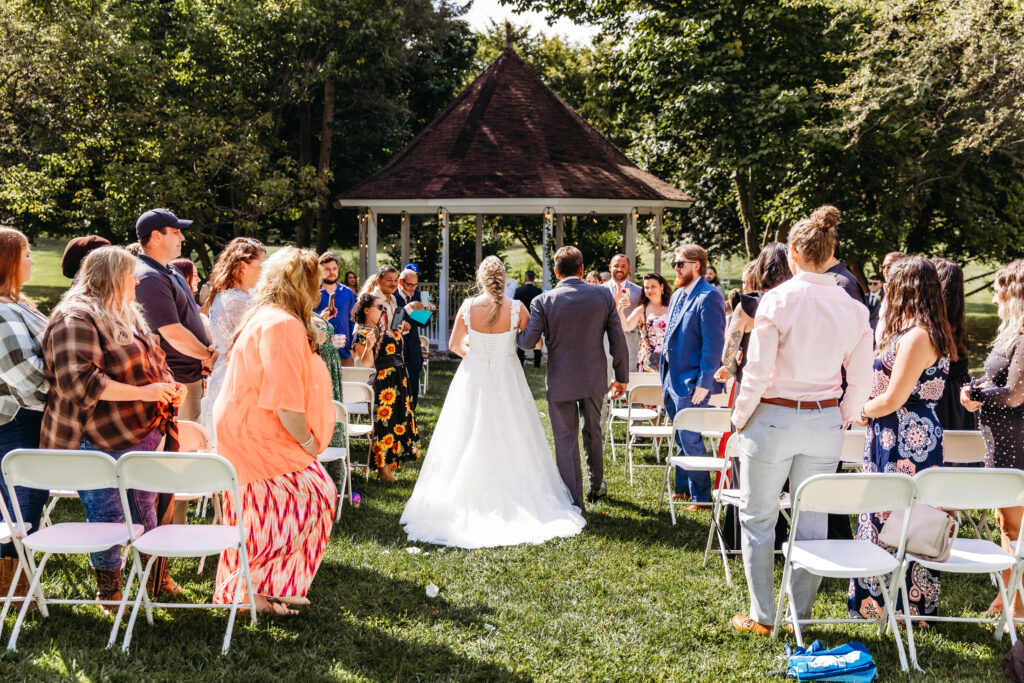 An outdoor wedding ceremony with guests standing and a couple facing each other near a gazebo. People are dressed in semi-formal attire on a sunny day.