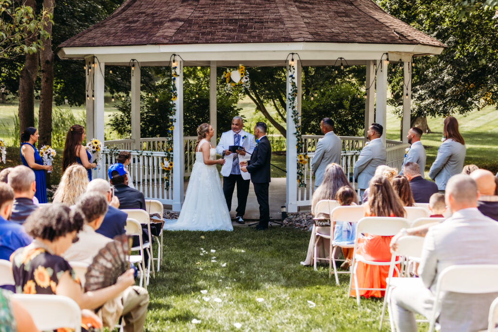 An outdoor wedding ceremony is underway with guests facing a couple exchanging vows under a gazebo adorned with sunflowers on a sunny day.