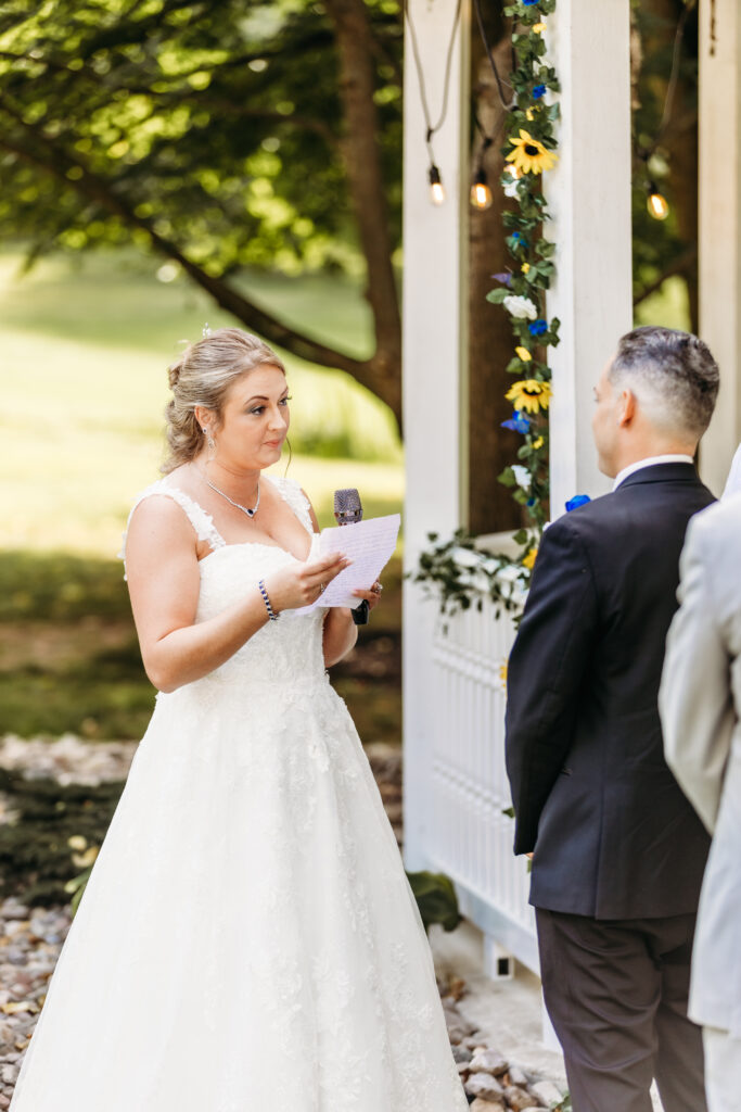 In an outdoor setting, a person in a wedding dress reads from a paper to another person facing them, surrounded by greenery and decor lights.